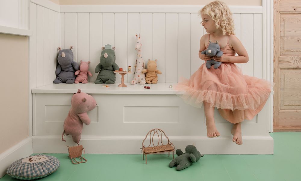 A young girl in a peach tutu dress sits on a white bench holding a soft toy, surrounded by Maileg stuffed animals arranged in playful positions on and around the bench.