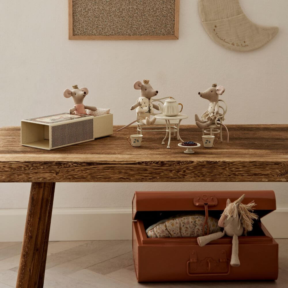 Three Maileg toy mice wearing crowns sit at a tiny tea table on a wooden surface, with miniature teacups and a teapot, while a toy horse rests in an open suitcase below.