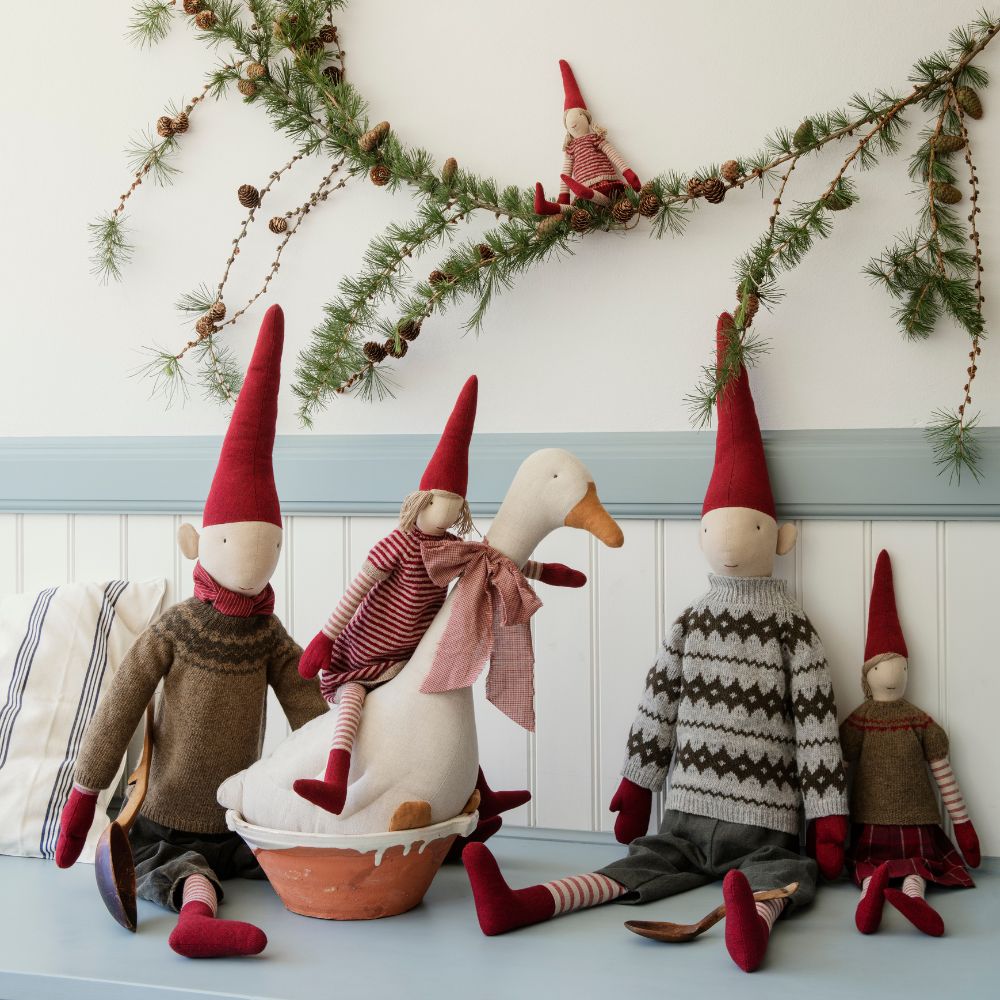 Maileg Christmas pixie dolls wearing red hats and festive jumpers sitting with a fabric goose under pine garland holiday decorations