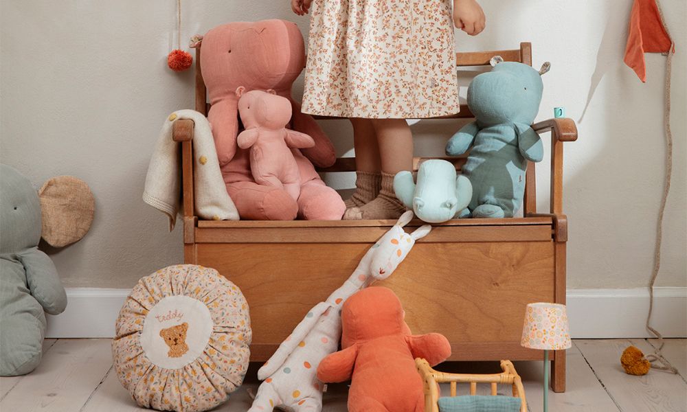 Child standing on a wooden bench surrounded by Maileg soft toys in pastel colours, including elephants, hippos, and rhinos in a cosy nursery setting.