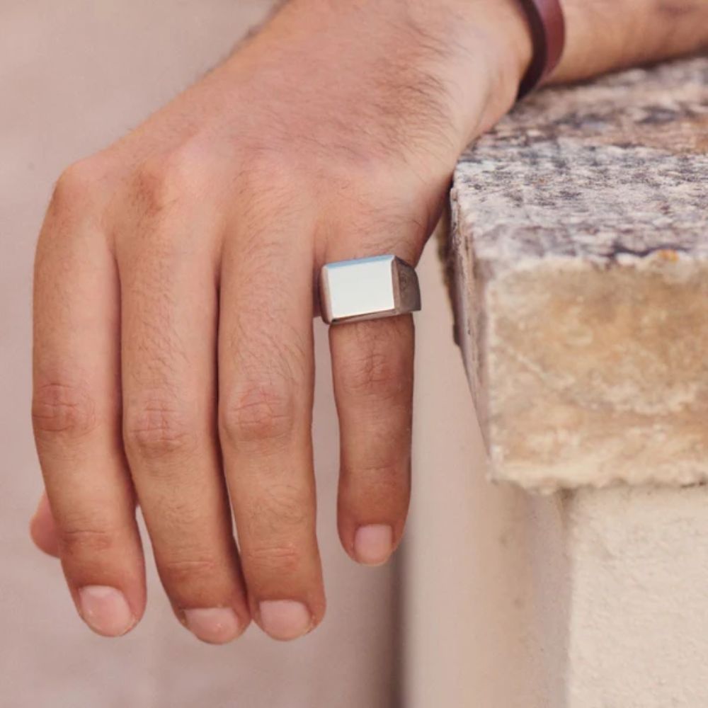 Hand wearing a silver square ring on a neutral background