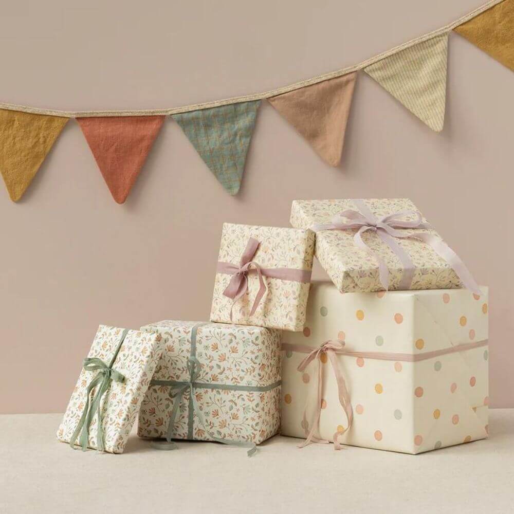 Stack of pastel Maileg gift boxes tied with ribbons and floral wrapping paper, with fabric bunting hanging in the background.