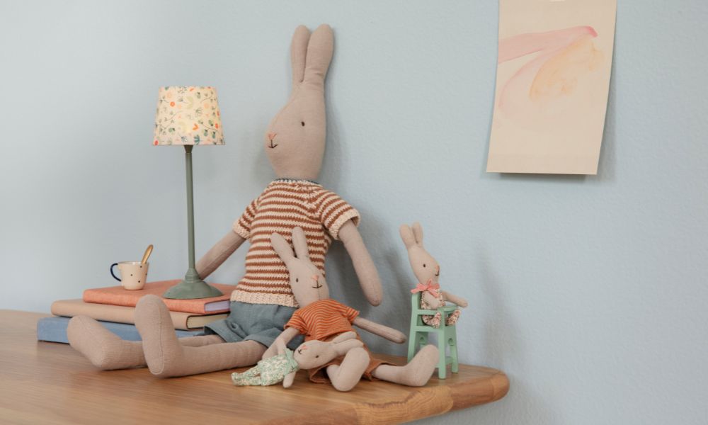 Maileg bunny soft toys in striped clothes sitting on a wooden table beside books and a small lamp in a cosy room setting.
