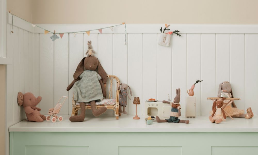 A group of Maileg soft toy animals, including rabbits and an elephant, are arranged in a charming miniature room with pastel furniture, bunting, and tiny accessories.
