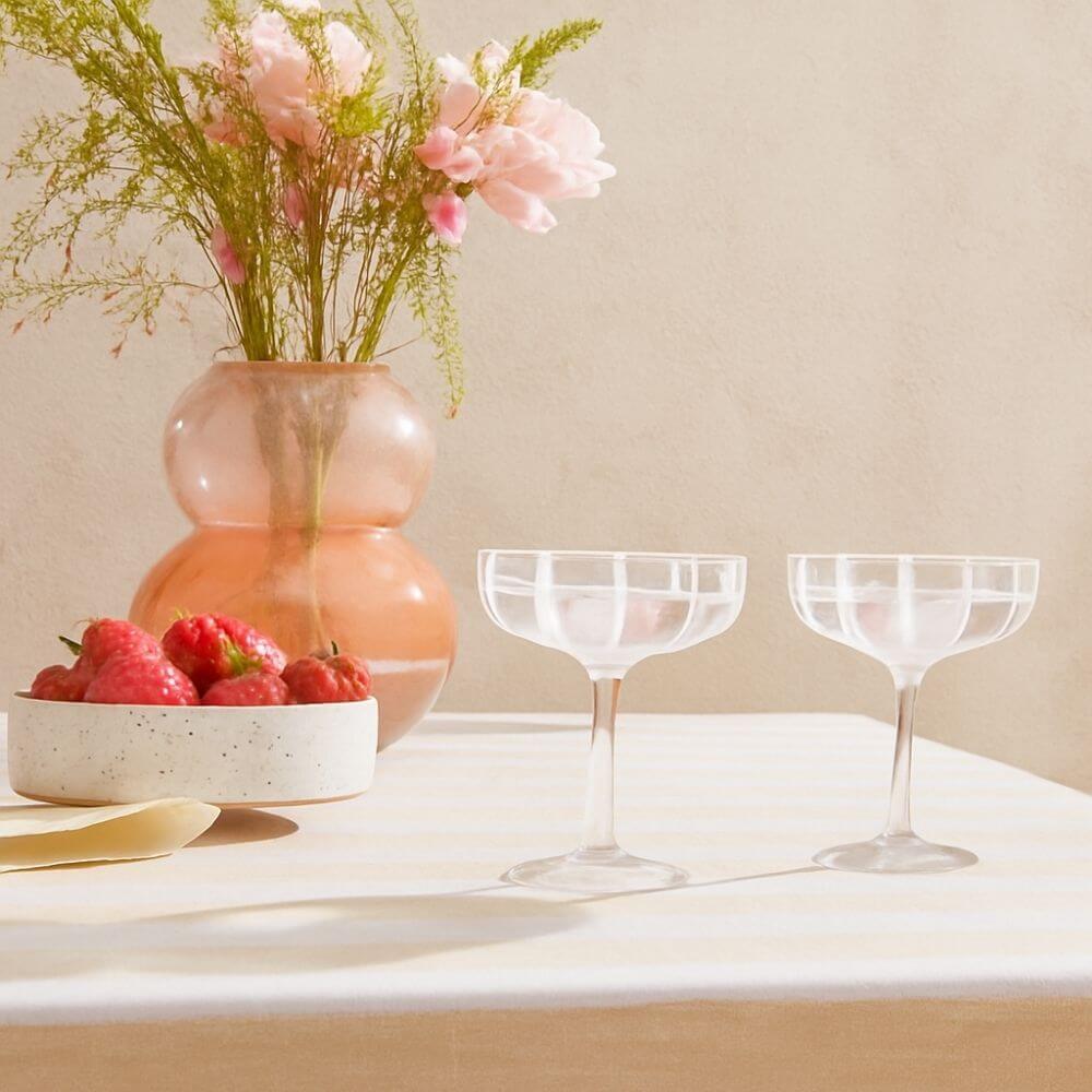 Decorative setting with a vase of flowers, bowl of strawberries, and two glasses on a light surface.